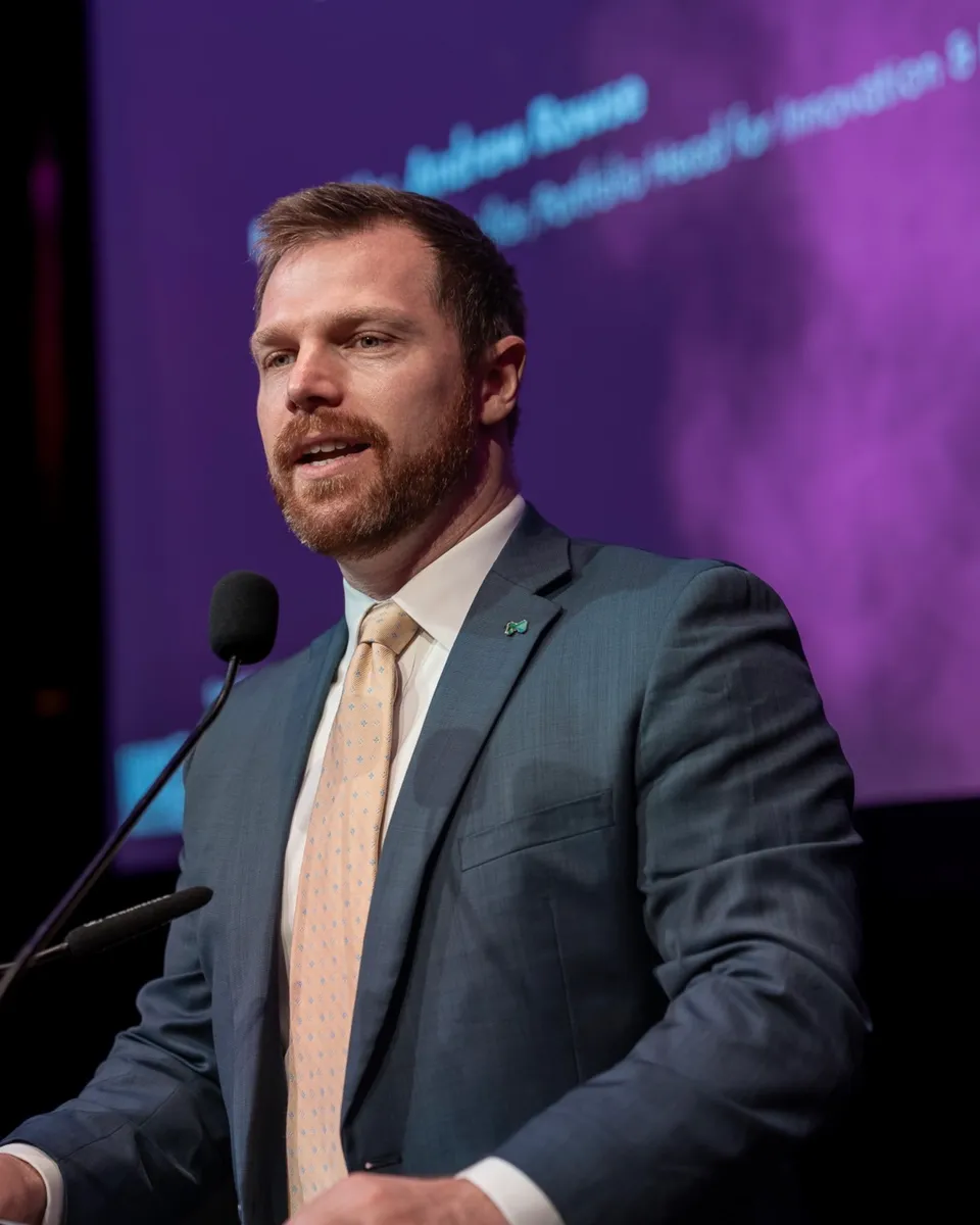 Andrew in a suit and tie, speaking at a lectern on stage