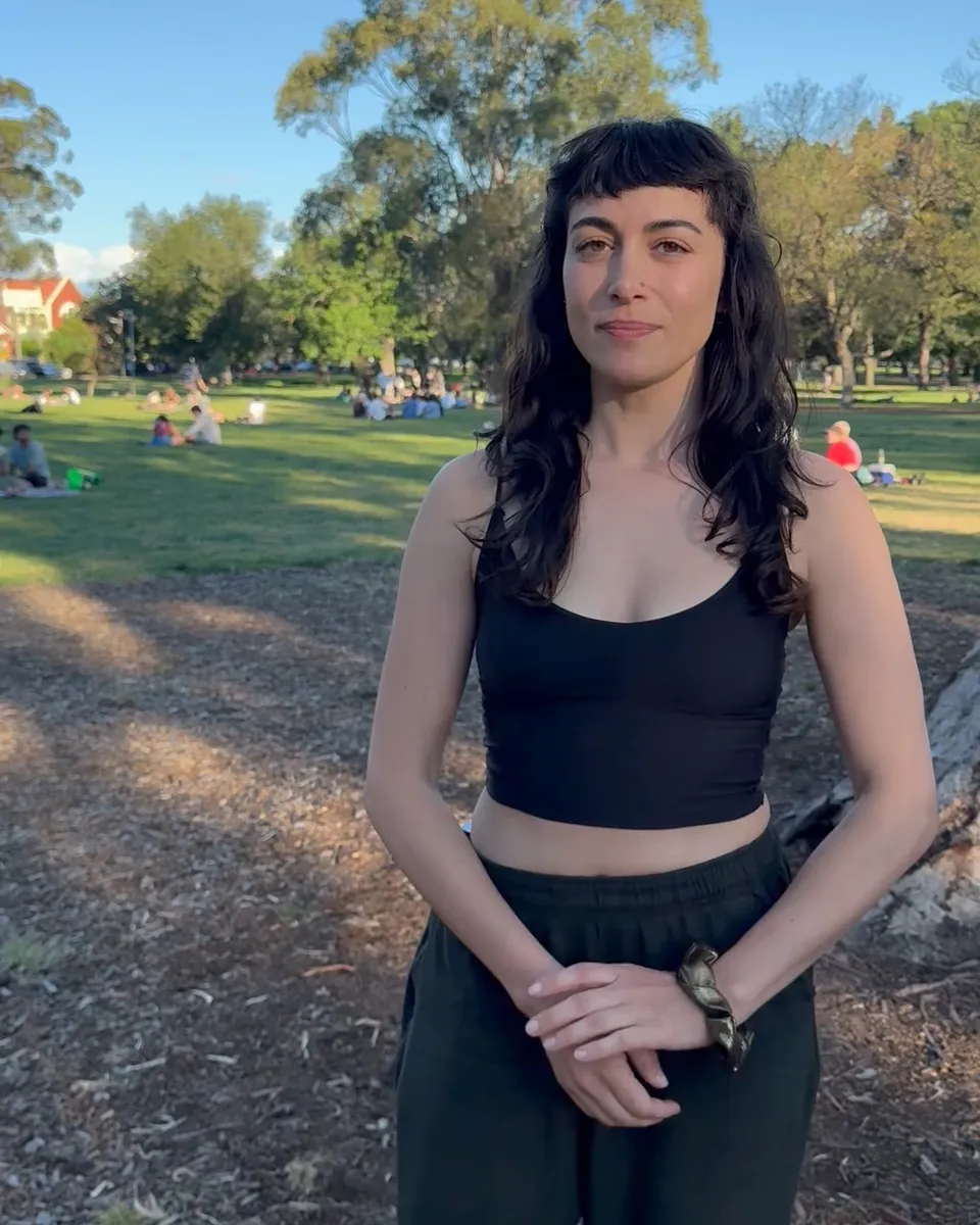 Sahar standing in a park with her hands clasped, smiling softly