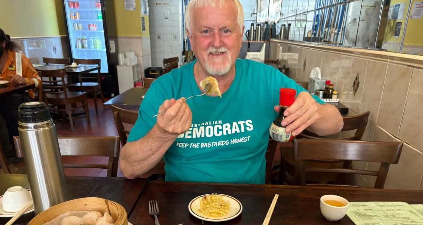 An Australian Democrats supporter in a teal 'Keep the Bastards Honest' T-shirt at a dumpling restaurant, holding up a dumpling on a fork and a bottle of soy sauce